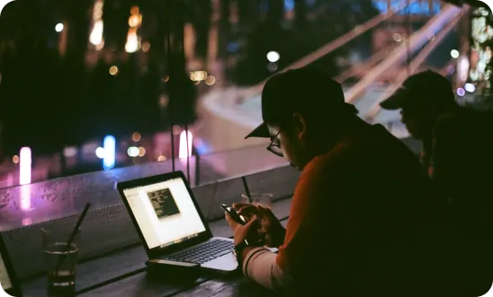 Person sitting at a table with a laptop and smartphone in a dimly lit cafe 
                            or lounge with colorful lights outside the window, 
                            creating a relaxed and focused atmosphere